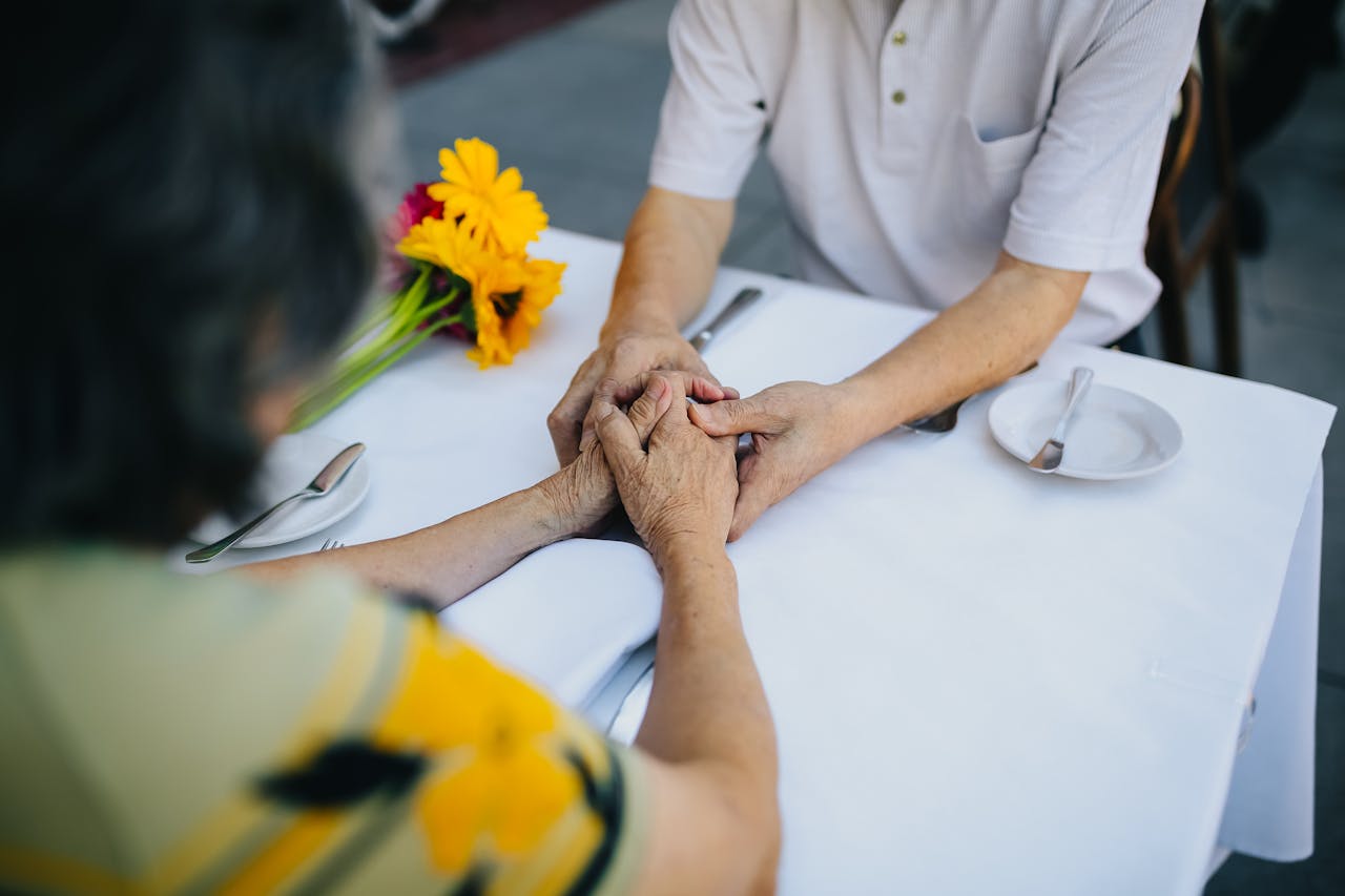 Elderly couple holding hands on a cafe table outdoors with a bouquet of colorful flowers.